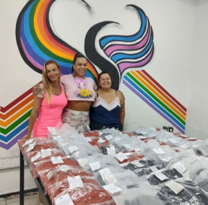 Três mulheres (Samara Arkcelio, Kamilly Santos e Juliana Alves) se posicionam atrás de uma mesa coberta de embalagens plásticas com roupas novas e panfletos. Atrás delas uma parede pintada com o logo da Casa Neon Cunha, bastante colorida.