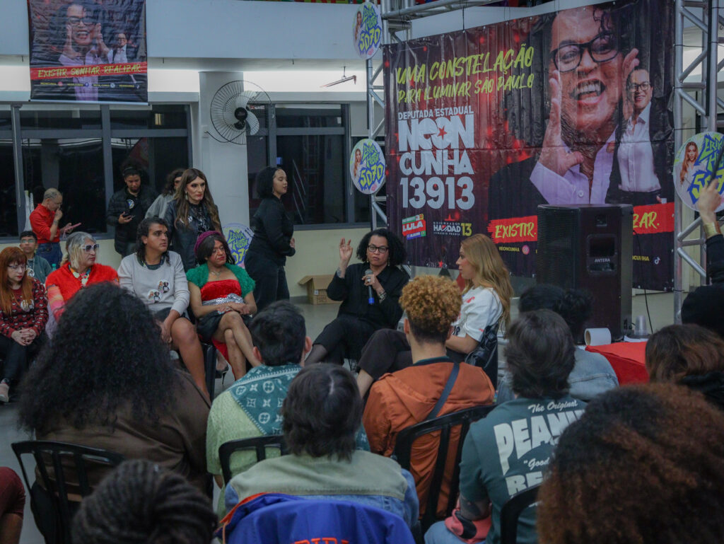 A imagem mostra uma reunião com dezenas de pessoas sentadas, de costas. À frente delas, no canto direito da foto, há um banner de cerca de 2m de altura com informações da candidatura de Neon Cunha. No centro, um pouco à frente do banner, Erika Hilton e Neon Cunha se encontram sentadas de frente para o público. Neon fala ao microfone.